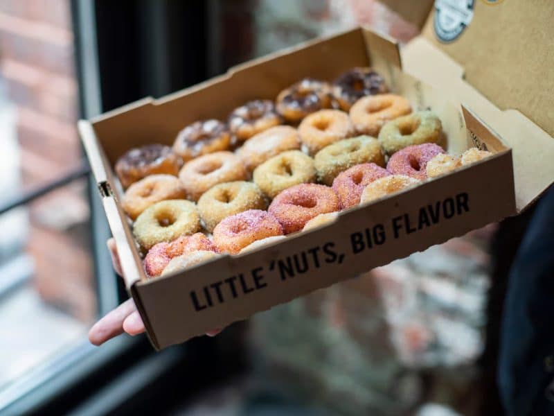 Box of assorted mini donuts with colorful toppings, labeled "LITTLE 'NUTS, BIG FLAVOR," held near a window, showcasing a variety of sweet treats perfect for a culinary adventure in Chelsea Market.