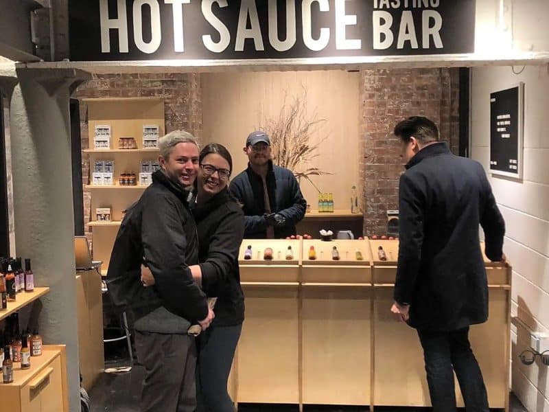 Couple smiling at Hot Sauce Tasting Bar in Chelsea Market, surrounded by various hot sauce bottles and a vendor, highlighting the culinary experience on the food tour.