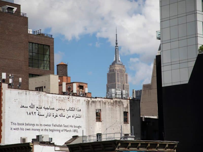 Empire State Building towering over urban rooftops with a historical mural in Arabic and English, representing the vibrant culture and history of New York City.