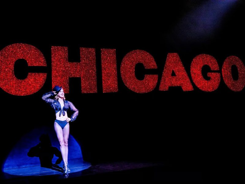 Dancer in black costume posing confidently in front of sparkling red "CHICAGO" sign, showcasing the vibrant energy of the Broadway show.