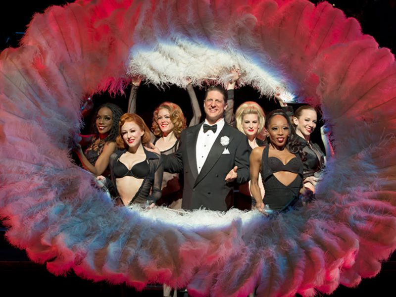 Cast of "Chicago" on stage, featuring a male lead in a tuxedo surrounded by female dancers in glamorous costumes, framed by a dramatic feather boa backdrop, showcasing the vibrant energy and theatrical flair of the Broadway show.