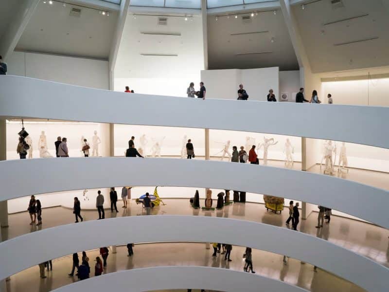 Interior view of the Solomon R. Guggenheim Museum showcasing visitors on spiraling ramps and art installations in the background, highlighting its architectural design by Frank Lloyd Wright and modern art exhibitions.