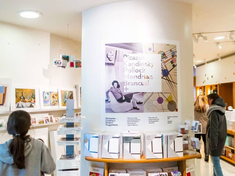 Visitors exploring the Guggenheim Museum gift shop featuring exhibition display on modern artists Picasso, Kandinsky, Pollock, Mondrian, and Brancusi, with various art books and merchandise available for purchase.