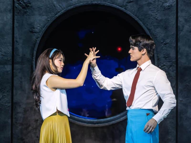 Darren Criss and Helen J Shen performing a romantic duet in "Maybe Happy Ending," featuring a cozy apartment setting and a circular window backdrop.
