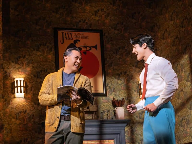 Two male performers smiling and engaging in a scene from the Broadway musical "Maybe Happy Ending," set in a cozy apartment with jazz-themed decor.