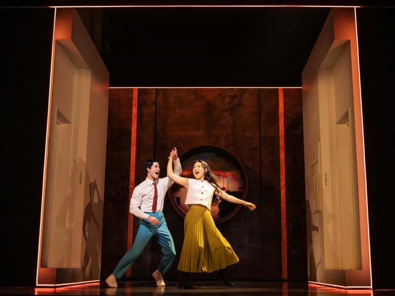 Darren Criss and Helen J Shen performing a lively dance in a cozy one-room apartment setting from the Broadway musical "Maybe Happy Ending."