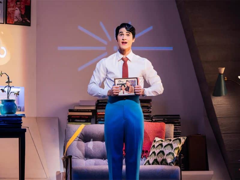 Darren Criss performing in "Maybe Happy Ending," holding a photo in a cozy apartment setting with jazz records and a plant.