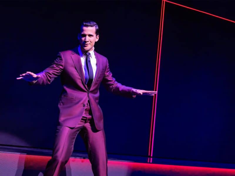 Darren Criss performing in a stylish burgundy suit on stage, promoting the Broadway musical "Maybe Happy Ending."