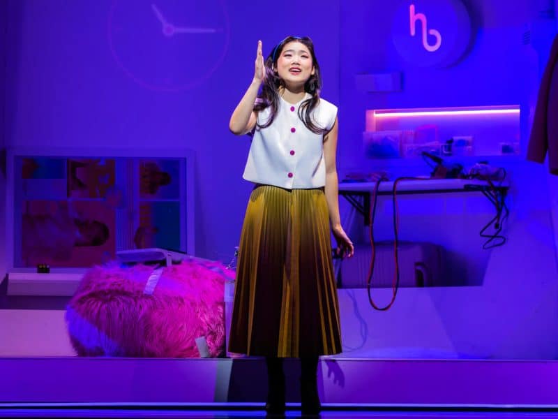 Helen J Shen performing in "Maybe Happy Ending," vibrant stage setting with purple lighting and pink decor, showcasing a cozy apartment atmosphere.