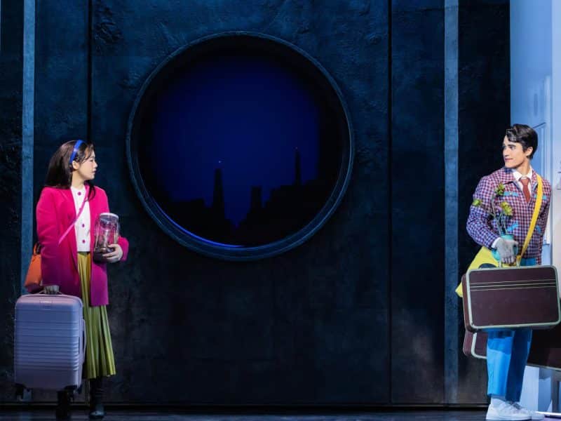 Girl in pink coat with suitcase and jar, standing next to a man with a suitcase, set in a cozy apartment scene from the Broadway musical "Maybe Happy Ending."