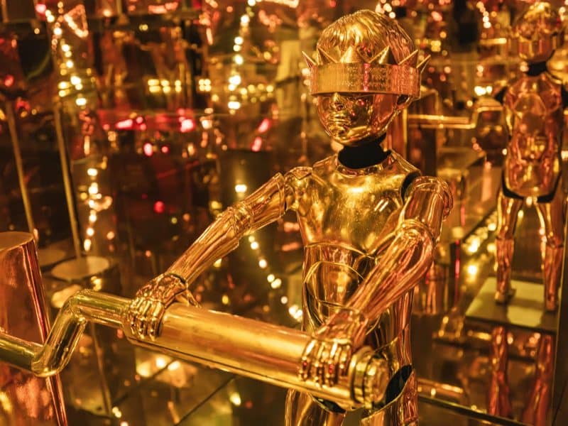 Golden robotic figure in an immersive exhibition room at Mercer Labs Museum of Art and Technology, surrounded by reflective surfaces and vibrant lights, showcasing innovative art and technology.