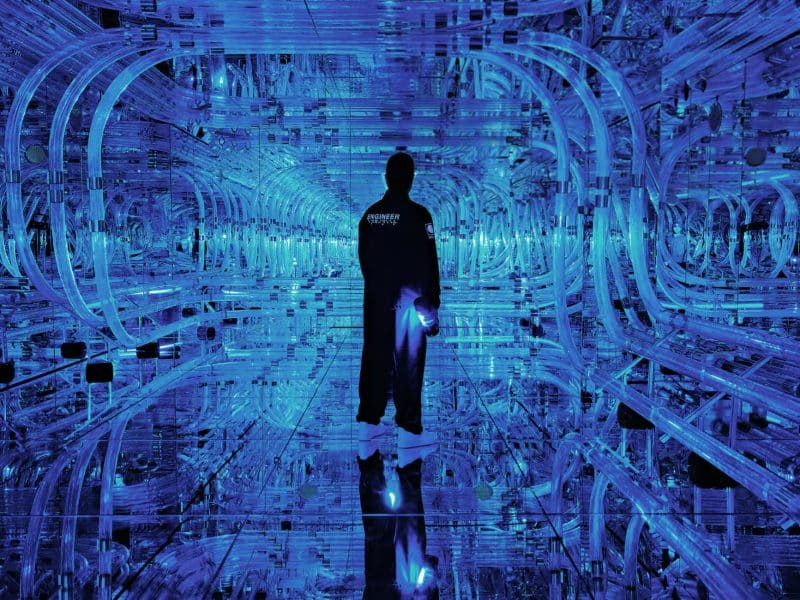 Person standing in a blue-lit immersive installation at Mercer Labs Museum of Art and Technology, surrounded by reflective surfaces and intricate light patterns.