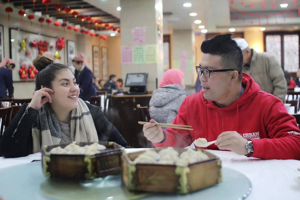 Smiling diners enjoying dumplings in a bustling Flushing restaurant, showcasing NYC's vibrant Chinatown culinary scene.