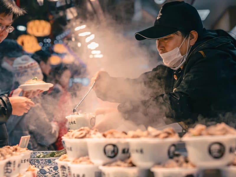 Vendor serving steaming bowls of traditional Asian cuisine at a bustling Flushing market, surrounded by eager customers and vibrant food stalls.