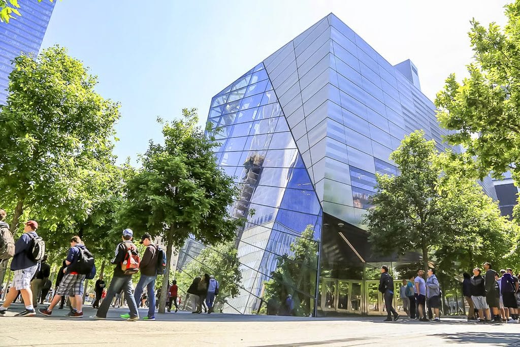 Visitors approaching the National September 11 Memorial Museum, featuring modern architecture and surrounded by trees, on a sunny day.