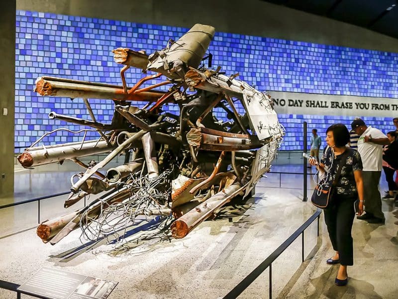 Artifact from 9/11 Memorial Museum, twisted metal beams and debris, with visitors reflecting, "No day shall erase you from the memory of time" inscription in background.