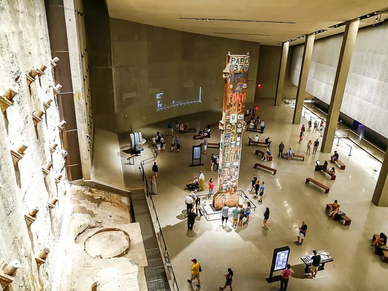 Interior view of the National September 11 Memorial Museum featuring the iconic "Survival Tree" sculpture, visitors exploring exhibits, and the textured wall commemorating the events of 9/11.