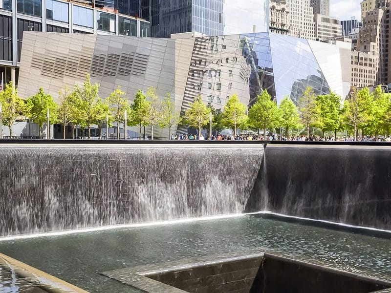 9/11 Memorial reflecting pool with cascading water, surrounded by trees and the National September 11 Memorial Museum's modern architecture in the background.