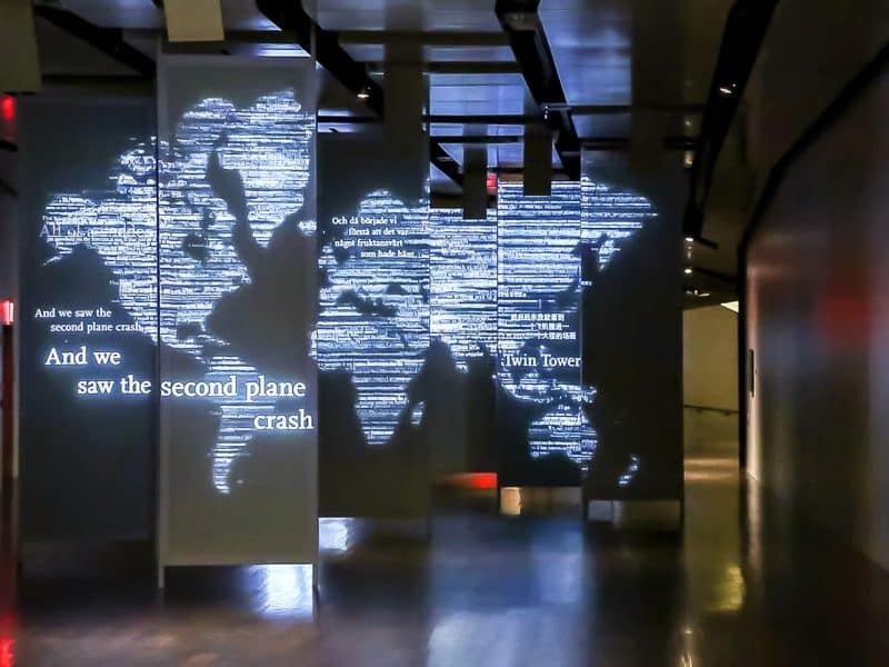 World map projection illuminated with text reflecting 9/11 experiences, including phrases like "And we saw the second plane crash" and references to the Twin Towers, within the National September 11 Memorial Museum.