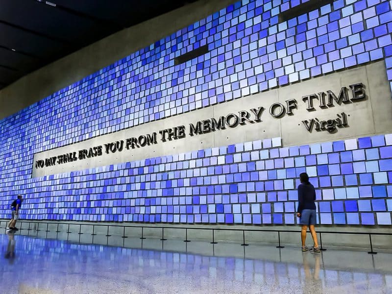 Visitor reflecting at the 9/11 Memorial Museum, featuring a wall with the inscription "No day shall erase you from the memory of time" by Virgil, surrounded by blue-tinted tiles.