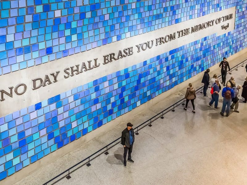 Interior of the 9/11 Museum featuring blue glass panels and the inscription "NO DAY SHALL ERASE YOU FROM THE MEMORY OF TIME," with visitors reflecting on the memorial's significance.