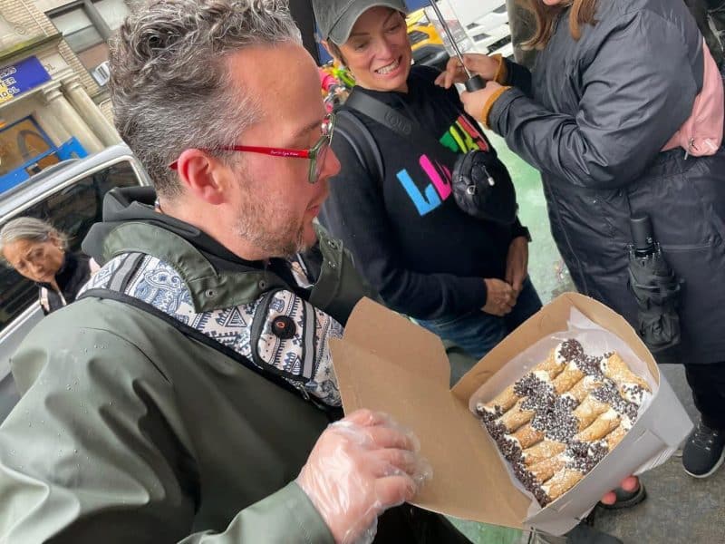 Man holding a box of cannoli with chocolate chips, surrounded by enthusiastic participants on the Original Greenwich Village Food & Culture Walking Tour.