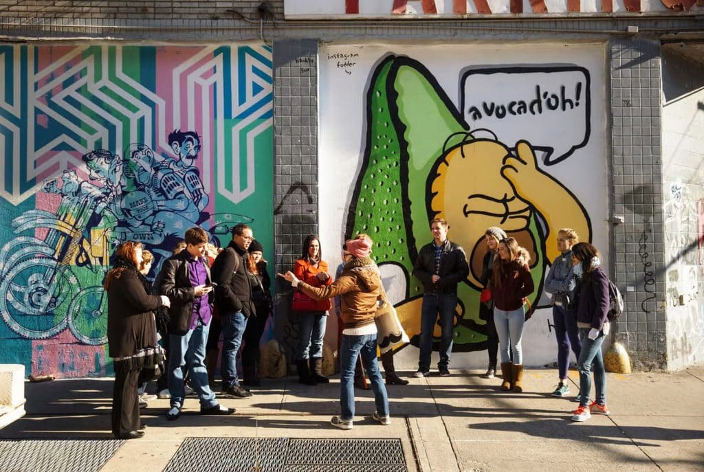 Group of tourists listening to a guide in front of colorful street art featuring an avocado character and vibrant murals, showcasing the cultural diversity of New York City's neighborhoods during a guided walking tour.