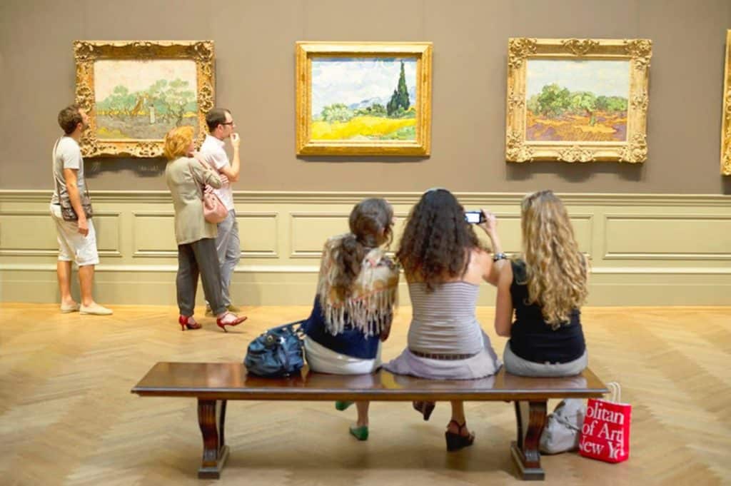 Visitors admiring artworks in the Metropolitan Museum of Art, featuring framed paintings on the wall and a group of people seated on a bench, capturing the experience on their phones.