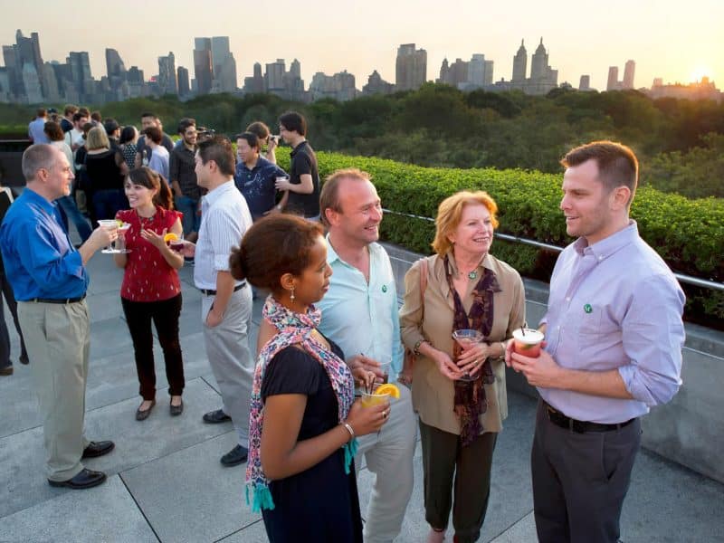 Group of diverse individuals enjoying drinks on a rooftop terrace with a view of the New York City skyline, socializing during a guided tour event at The Met Museum.