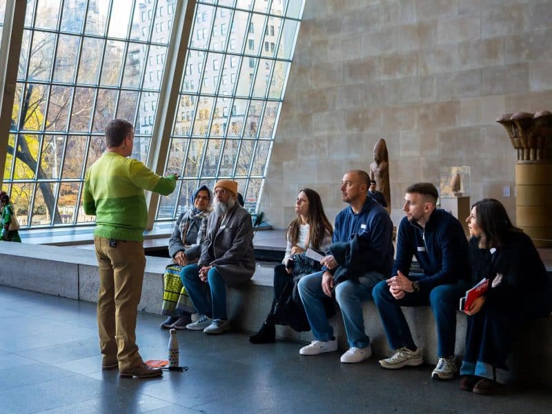 Group of visitors listening to a guide in the Great Hall of the Metropolitan Museum of Art during a guided tour, featuring large glass windows and sculptures in the background.