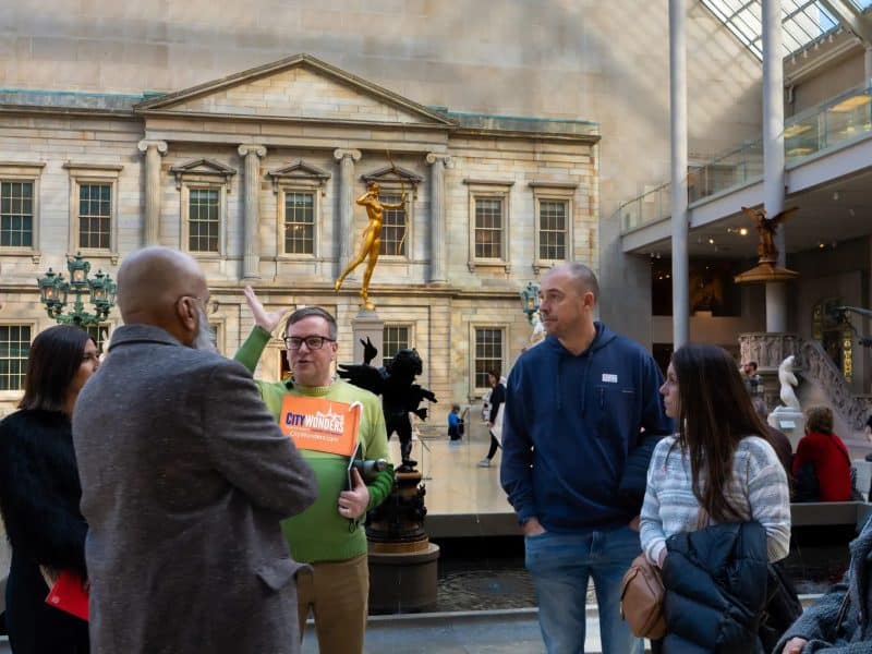 Group of tourists engaging in a guided tour at the Metropolitan Museum of Art, with a tour guide holding a "City Wonders" sign, surrounded by iconic sculptures and the museum's Great Hall backdrop.