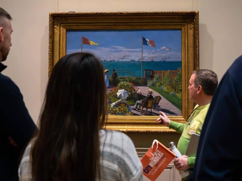 Visitors observing a painting during a guided tour at the Metropolitan Museum of Art, featuring a vibrant seaside scene with flags and figures, led by an expert guide.