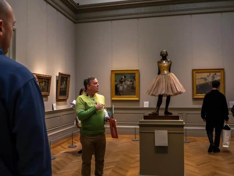 Guided tour at The Met Museum featuring a statue of a ballet dancer, with a guide in a green sweater discussing art pieces in the background, surrounded by iconic paintings.