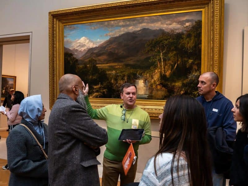 Group of visitors listening to a guide in the Metropolitan Museum of Art, discussing a landscape painting, with the Great Hall's architecture in the background.