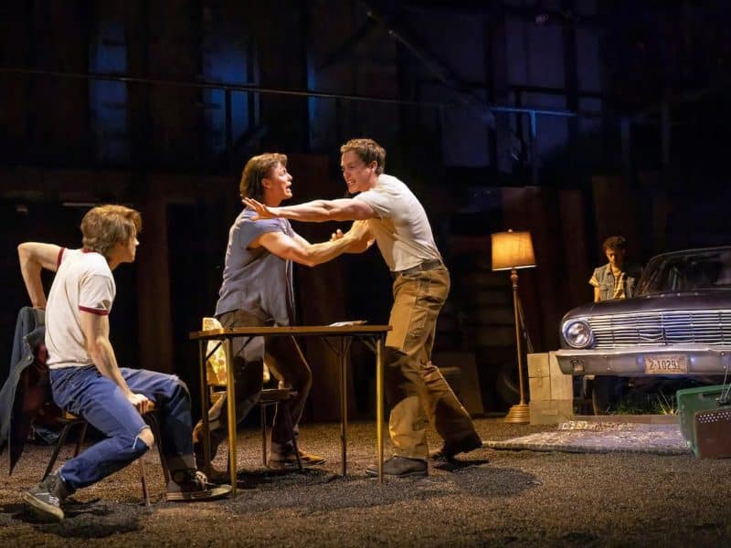 Scene from "The Outsiders" musical depicting a tense moment between characters at a table, with two male actors in conflict, a seated character observing, and a vintage car in the background, set in a 1967 Tulsa, Oklahoma environment.
