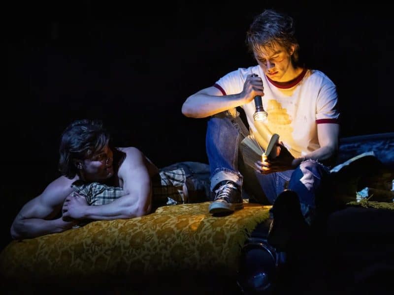 Two young men on a yellow bed, one reading a book with a flashlight, the other lounging beside him, evoking themes of friendship and self-discovery from "The Outsiders" musical set in 1967 Tulsa.