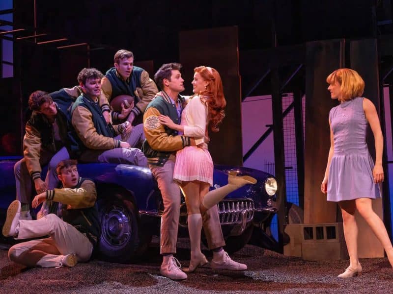 Cast of "The Outsiders" musical performing on stage, featuring characters in 1960s attire, with a vintage car backdrop, highlighting themes of friendship and rivalry.