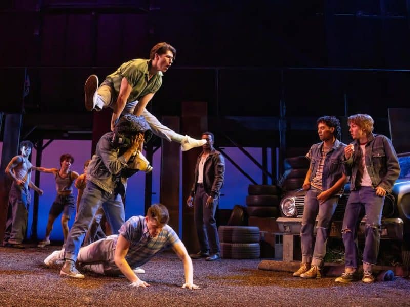 Cast of "The Outsiders" musical performing dynamic choreography on stage, showcasing themes of friendship and resilience amidst a vibrant Tulsa backdrop.
