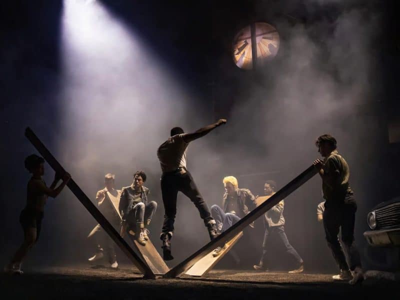 Group of young performers in dynamic poses on a stage, depicting scenes from "The Outsiders" musical, with dramatic lighting and a vintage car in the background.