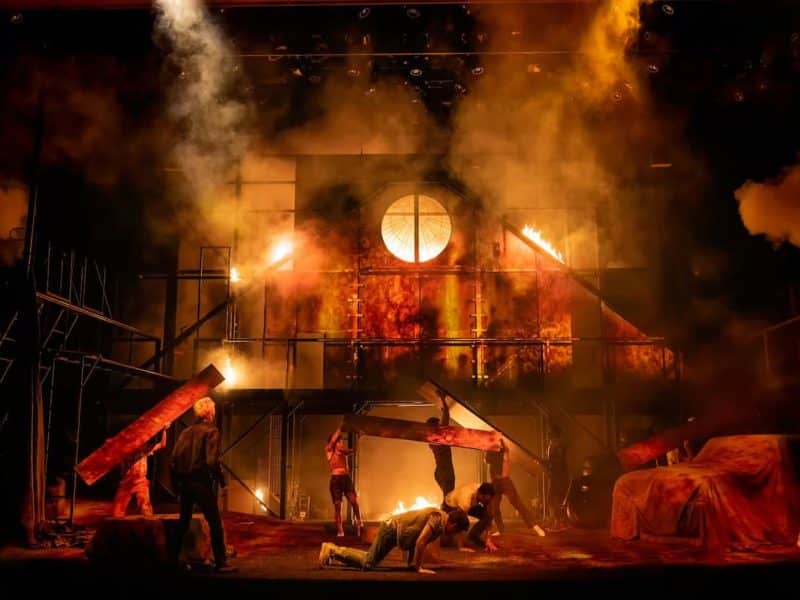 Theatrical scene from "The Outsiders" musical, featuring dramatic lighting, smoke, and actors in dynamic poses amid a fiery backdrop, representing themes of struggle and belonging.
