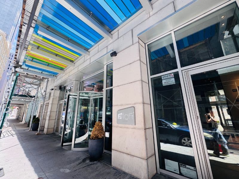 Aloft Manhattan Downtown hotel entrance with modern glass doors, colorful awning, and stylish architecture, located near Fulton Street subway, highlighting pet-friendly and accessible features.