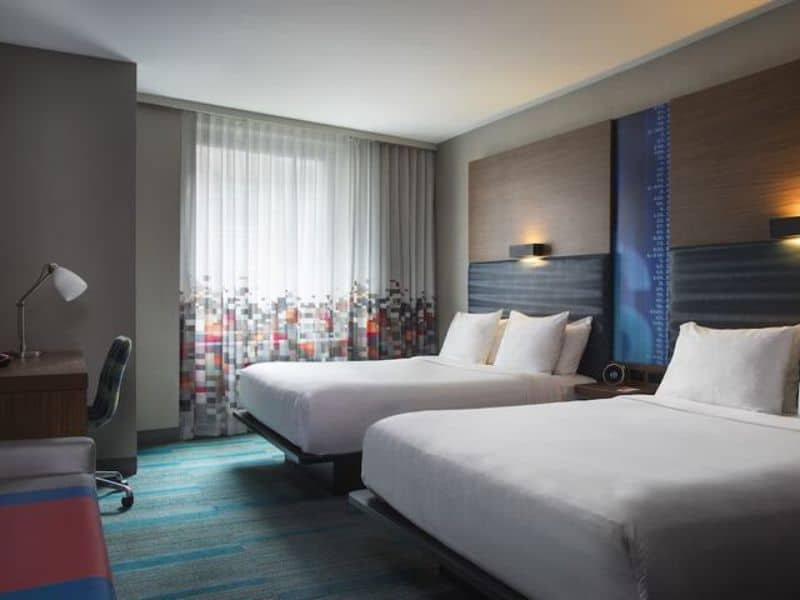 Modern hotel room at Aloft Manhattan Downtown featuring two queen beds, ergonomic desk, and colorful window treatments, showcasing stylish accommodations ideal for business and leisure travelers.