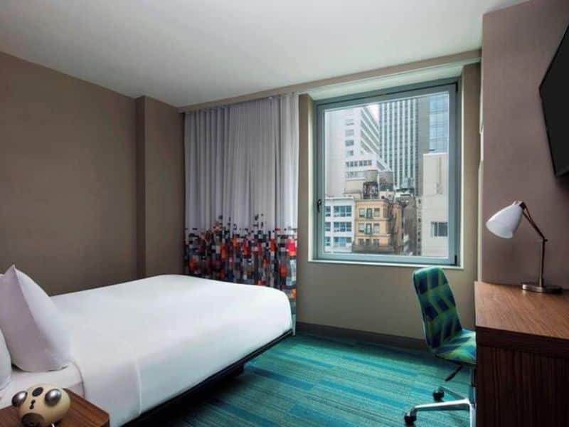Modern hotel room at Aloft Manhattan Downtown, featuring a large window with city views, colorful curtains, a comfortable bed, ergonomic desk chair, and stylish decor.