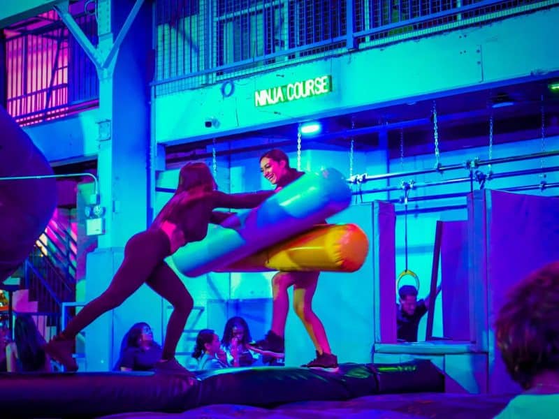 Two participants engaging in a playful duel on inflatable obstacles in the vibrant indoor ninja course at Area 53 NYC Adventure Park, surrounded by colorful lighting and spectators.