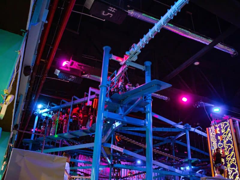 Indoor adventure park equipment with colorful lighting, featuring a zip line course and rock climbing wall at Area 53 NYC Adventure Park.