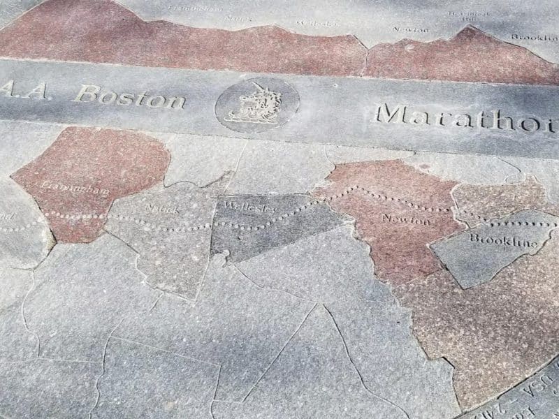 Map of Boston Marathon route showcasing neighborhoods including Newton, Wellesley, and Brookline, engraved in stone with "A.A. Boston" and "Marathon" inscriptions.