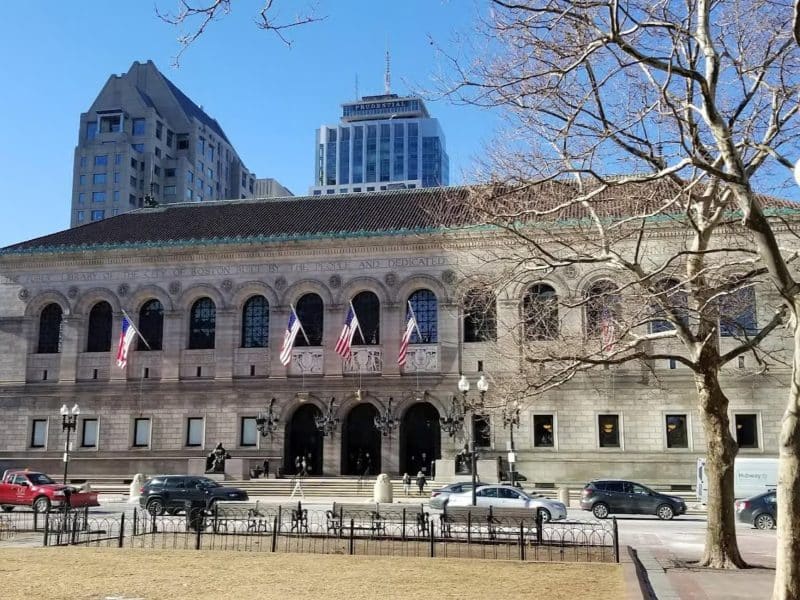 Boston Public Library's historic fa&ccedil;ade with American flags, surrounded by modern buildings and trees, showcasing the architectural blend of Boston's rich history and contemporary urban landscape.