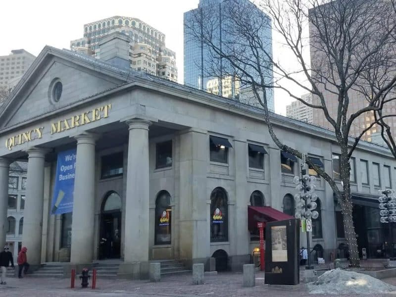 Quincy Market building in Boston with historic architecture, featuring a "We're Open for Business" banner, surrounded by modern skyscrapers and winter trees.