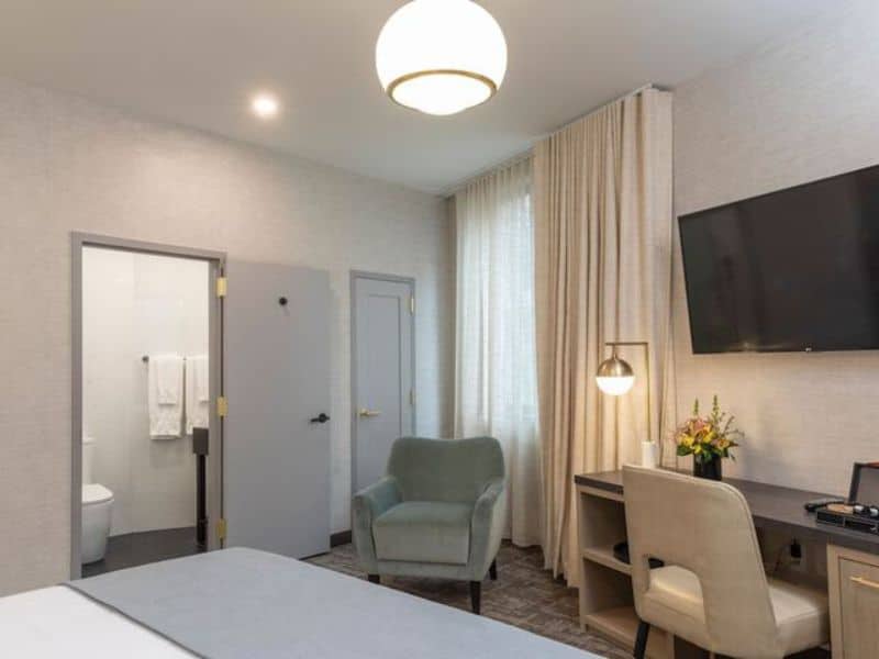 Elegant hotel room at Broadway Plaza Hotel featuring a cozy green chair, flat-screen TV, and modern decor, with a bathroom door visible and natural light from large windows.