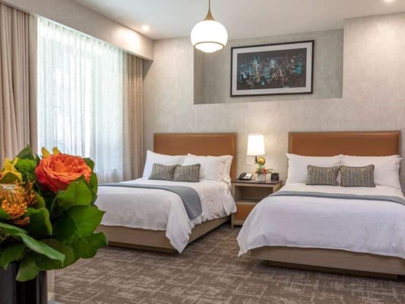 Stylish hotel room at Broadway Plaza Hotel featuring two beds, contemporary decor, flat-screen TV, and a floral arrangement, highlighting modern amenities in the Nomad district of NYC.